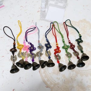 May include: A collection of eight decorative charms with bells, coins, and knotted cords in various colors. Each charm features a small bell, a coin with a square hole, and a decorative knot. The cords are black, yellow, pink, purple, gray, blue, red, green, and dark green.