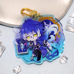 May include: A clear acrylic charm featuring a cartoon character with blue hair and yellow eyes. The charm has a gold-colored clasp and a blue and teal background with a decorative border. The character is holding a book and a lantern.