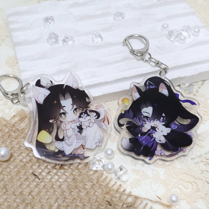 May include: Two clear acrylic keychains featuring anime-style characters with black hair and animal ears. One keychain shows a character in white, holding a small dog. The other shows a character in purple, holding a white cat. Both have silver key rings.