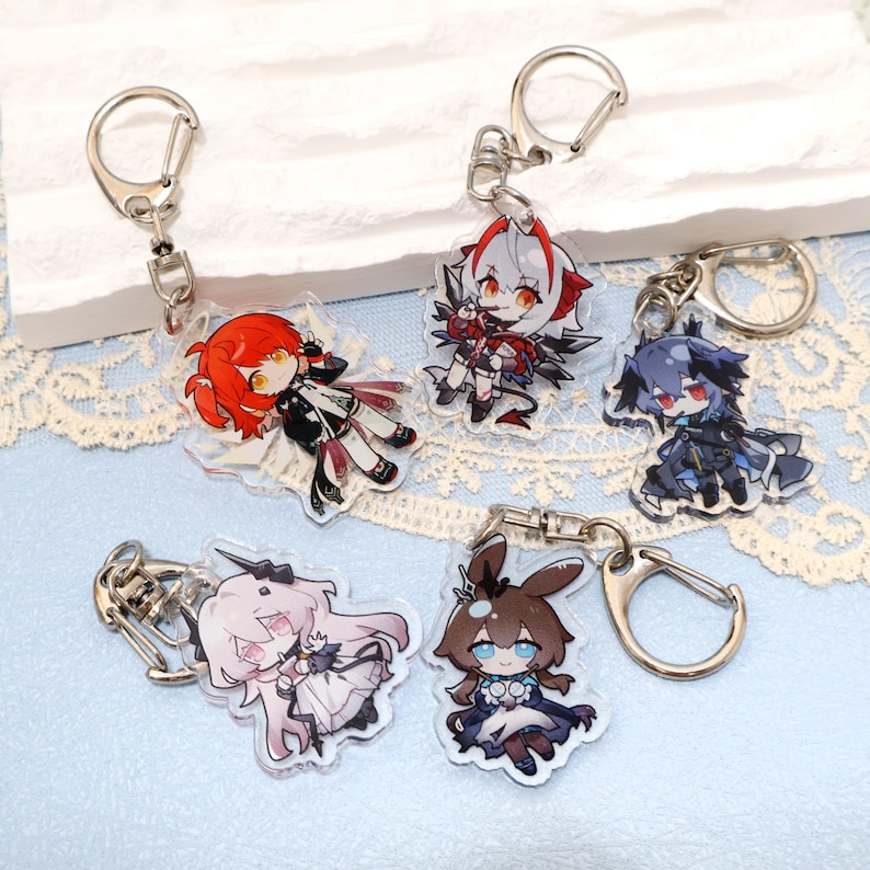 May include: Five acrylic keychains featuring anime-style characters with vibrant colors. Each keychain has a silver-colored clasp. The characters have detailed designs, with red, white, black, and blue color schemes. The keychains are on a light blue surface.