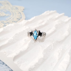May include: A silver ring with a light blue, diamond-shaped gemstone at the center. The ring features black bat-wing designs on either side of the gemstone. The band has a detailed, openwork pattern. The ring is displayed on a white, textured surface.