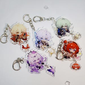 Könnte beinhalten: Eine Sammlung von Acryl-Schlüsselanhängern mit Charakterdesigns im Anime-Stil. Jeder Schlüsselanhänger hat einen silberfarbenen Metallverschluss und eine Kette. Die Charaktere sind in leuchtenden Farben gehalten, mit Details wie Haaren, Kleidung und Accessoires.