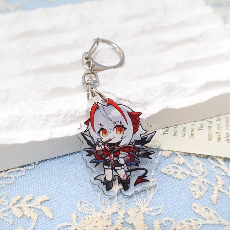 May include: A clear acrylic keychain featuring a chibi anime character with white hair, red accents, and black wings. The character is holding a small book and has a devil tail. The keychain has a silver metal clasp.