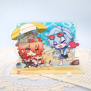 以下が含まれることがあります： ビーチにいる2人のアニメ風キャラクターが描かれた透明なアクリルスタンド。一人は黄色のパラソルの下にいて、もう一人は赤い魚を持っています。背景には砂浜と海のあるビーチの風景が描かれています。