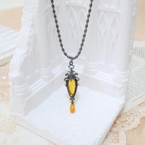 May include: A pendant necklace with a dark metal chain and ornate pendant. The pendant features a yellow, teardrop-shaped stone with a decorative metal frame. The necklace is displayed against a white, architectural backdrop.