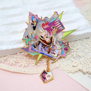 May include: An ornate enamel pin depicting an anime-style character within a starburst design. The pin includes a gold chain with two small, dangling card-shaped charms. The colour scheme features purple, pink, green, and gold.