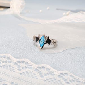 May include: A silver ring featuring a light blue, diamond-shaped gemstone. The ring has black accents and a unique design. The ring is displayed on a light blue surface with white lace.