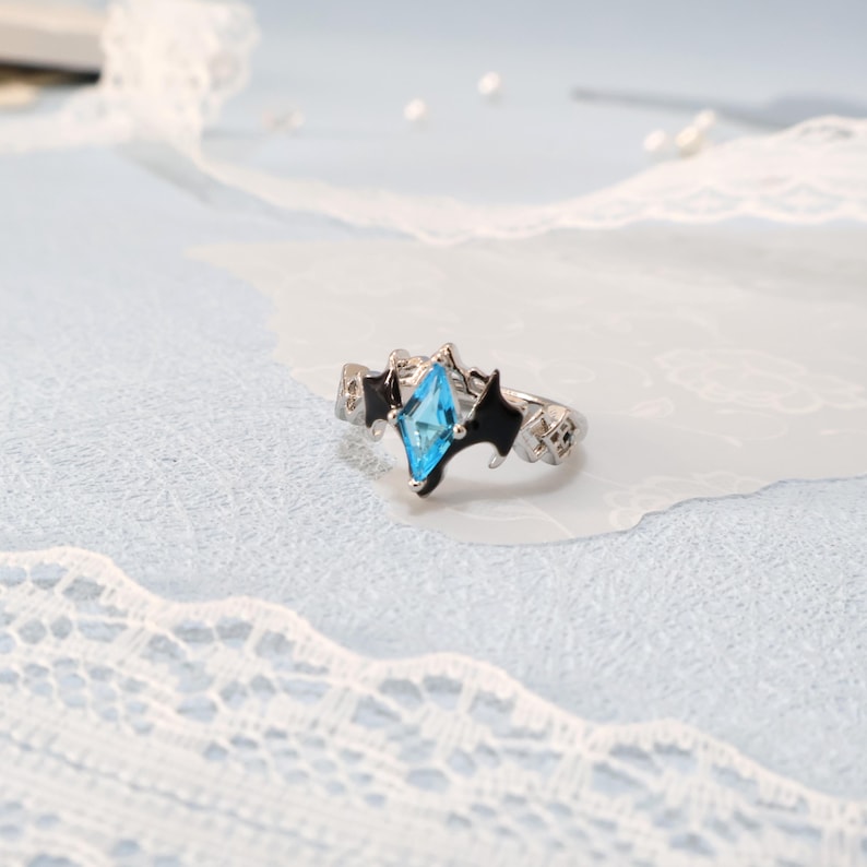 May include: A silver ring featuring a light blue, diamond-shaped gemstone. The ring has black accents and a unique design. The ring is displayed on a light blue surface with white lace.