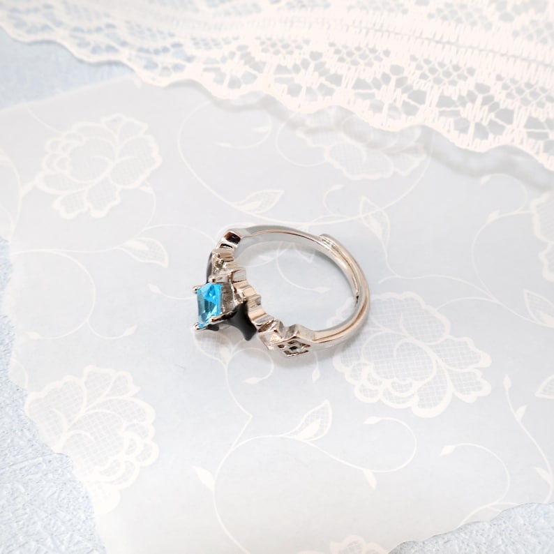 May include: A silver-toned ring with an adjustable band. The ring features a rectangular, light blue gemstone. The band has decorative cutouts and is set against a floral patterned background.