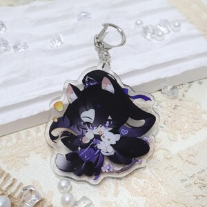 May include: A clear acrylic keychain featuring a cartoon character with black hair, purple accents, and a white cat. The keychain has a silver clasp and a floral design. The character is holding a small white cat.