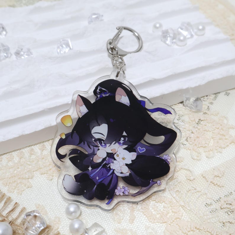 May include: A clear acrylic keychain featuring a cartoon character with black hair, purple accents, and a white cat. The keychain has a silver clasp and a floral design. The character is holding a small white cat.