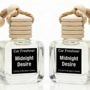 May include: Two clear glass car air fresheners with wooden tops and beige rope hangers. Each bottle has a black label with the words "Car Freshner Midnight Desire". The bottles are filled with a clear liquid.
