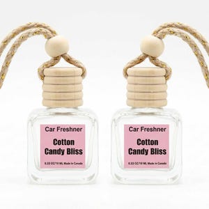 May include: Two clear glass car air fresheners with wooden caps and beige rope hangers. Each has a pink label with the text "Car Freshner Cotton Candy Bliss". The bottles are 0.33 oz/10 ml.
