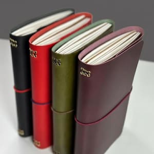 May include: Four leather journals in black, red, olive green, and burgundy. Each journal has a matching elastic closure and gold-colored corner accents. The pages are white and the edges of the pages are colored to match the cover.