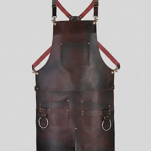 Może przedstawiać: Ciemnobrązowy skórzany fartuch z kieszenią na piersi i dwiema dolnymi kieszeniami. Fartuch posiada regulowane paski z mosiężnym osprzętem i czerwonymi akcentami. Do dolnych kieszeni przymocowane są dwa duże srebrne pierścienie.
