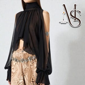 May include: Black sheer top with long sleeves and a high neck. The top has a cape-like design with open shoulders. The model is wearing a silver arm cuff and leopard print pants. The logo "MARRIANNE STYLES" is in the upper right corner.