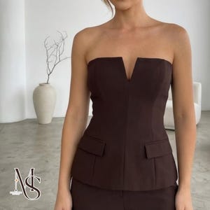 May include: A dark brown strapless top with a V-neck and two front pockets. The top is part of a suit, indicating formal wear. The fabric looks smooth and tailored. The outfit is suitable for business or evening wear.