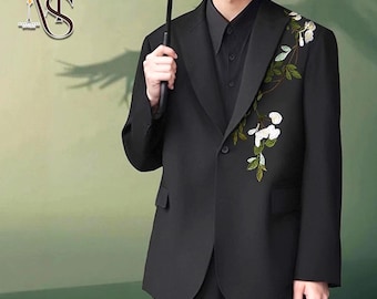May include: A black blazer with floral embroidery on the lapel. The blazer has a single-button closure and a classic notched collar. The floral design features white flowers and green leaves. The model is wearing a black shirt underneath.