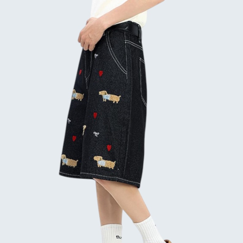 May include: Black denim shorts with white stitching and embroidered dachshund dogs and red hearts. The shorts have a high waist and a belt. The model is wearing white socks.