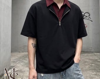 May include: A black short-sleeved shirt featuring a half-zip front closure and a contrasting dark red collared neckline. The garment is worn with light blue distressed denim jeans. The shirt has a clean, solid black fabric construction.