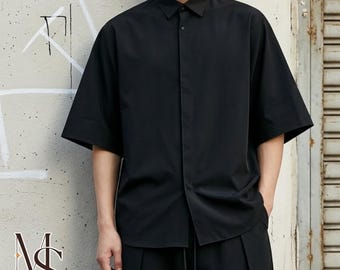 May include: A black short-sleeved button-down shirt with a pointed collar and a concealed front placket. The shirt features a loose, boxy fit with wide sleeves and a straight hemline, constructed from a smooth, solid-colored fabric.