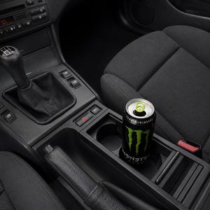 May include: A black and green Monster Energy drink can in a car's cup holder. The can has a black background with a green claw mark design and the Monster Energy logo. The car interior is black, with a manual gear shift visible.