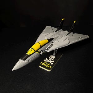Könnte beinhalten: Graues und gelbes F-14 Tomcat Modellflugzeug mit schwarzen Akzenten. Das Modell wird auf einem schwarzen Ständer mit dem Text "F-14 TOMCAT" und einer Totenkopf-Grafik präsentiert. Das Modell hat die Nummer "94" an der Seite.