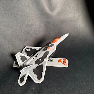 May include: A model airplane with a gray, black, and orange camouflage design. The toy airplane has a white base with a red and white logo. The wings and body of the plane are detailed with black and gray patterns.