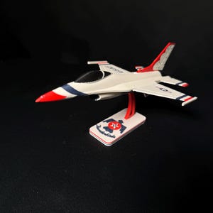 Könnte beinhalten: Ein weißes Modellflugzeug mit roten und blauen Akzenten, einschließlich Streifen und Flügelspitzen. Das Modell ist auf einem Ständer mit dem Wort "Thunderbirds" und einem Logo montiert. Das Flugzeug hat eine rote Nase und ein schwarzes Cockpit.