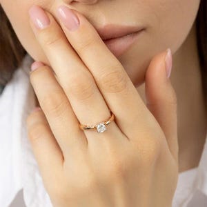 Puede incluir: Un anillo de oro con una piedra preciosa clara y redonda en el centro. El anillo está en el dedo de una persona, cerca de su rostro. El anillo tiene un diseño simple y elegante.
