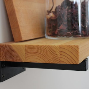 May include: A wooden shelf with a natural finish, supported by black metal brackets. A glass jar filled with dried herbs sits on the shelf, alongside a wooden cutting board. The shelf is mounted on a white wall.