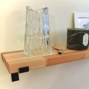 May include: A wooden shelf with black metal brackets supports a clear glass vase, a black marble-effect candle, a framed picture, and a small decorative box. The shelf is made of light-coloured wood.