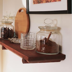 May include: A dark stained wooden shelf displays glass jars filled with dried herbs and spices. A wooden chopping board and a decorative glass are also on the shelf. The shelf is mounted on a white wall.