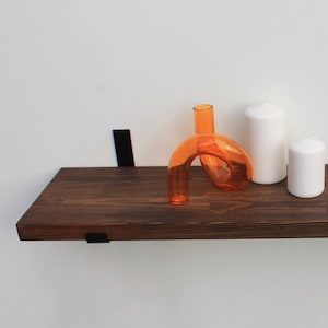 May include: A dark brown wooden floating shelf with black metal brackets, holding an orange glass sculpture and two white candles. The shelf is mounted on a white wall, creating a minimalist display.