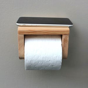 May include: A wooden toilet paper holder with a shelf for a phone. The holder is made of light-colored wood and has a roll of white toilet paper. A black smartphone rests on top of the holder.