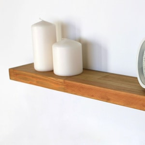 May include: A wooden shelf with a light brown finish, holding two white pillar candles and a white alarm clock. The clock has a round face with black hands and a silver handle. The shelf is mounted on a white wall.