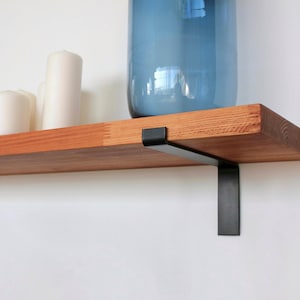 May include: A wooden floating shelf with a natural finish, supported by black metal brackets. The shelf holds white candles and a large blue glass vase. The shelf is a stylish storage solution, approx. 60cm long.