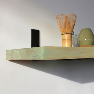 May include: A light green wooden shelf displaying a bamboo whisk, a small green ceramic vessel, and a green ceramic bowl. The shelf is mounted on a white wall with a black metal bracket.