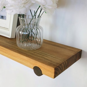 May include: A wooden floating shelf with a natural finish, holding a clear glass vase with white flowers and a framed picture. The shelf is mounted on a white wall and has a dark brown wooden peg.