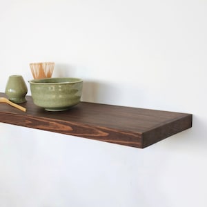 May include: A dark brown wooden floating shelf with a matcha bowl, whisk, spoon, and a small ceramic container. The shelf is mounted on a white wall. The items are green and natural wood.
