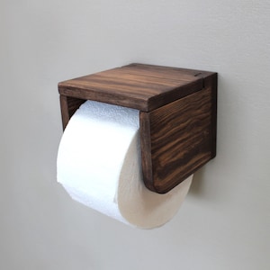 Può includere: Un portarotolo di carta igienica in legno marrone scuro, montato su una parete grigio chiaro. Contiene un rotolo di carta igienica bianca. Il design è semplice e moderno, con un focus sul legno.