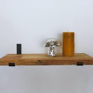 May include: A wooden shelf with black metal supports, displaying a silver disco mushroom ornament and a large, golden-yellow pillar candle. The shelf is mounted on a white wall, creating a minimalist display.