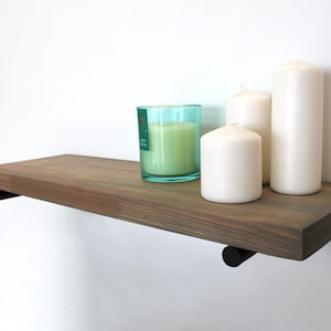 May include: A wooden floating shelf with black metal brackets, displaying a teal glass candle holder with a green candle and three white pillar candles. The shelf is mounted on a white wall.