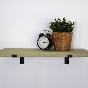 May include: A wooden floating shelf with black metal brackets, holding a black alarm clock and a potted plant in a woven basket. The shelf is painted a light green colour.