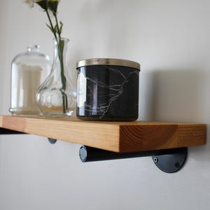 May include: A wooden floating shelf with black metal brackets, holding a black marble-patterned candle jar, a clear glass vase with flowers, and a glass jar. The shelf is made of light-coloured wood.