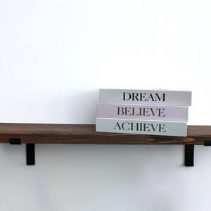 May include: A dark brown wooden floating shelf with black metal brackets, holding three books. The books are white with the words "DREAM", "BELIEVE", and "ACHIEVE" printed on the spines. The shelf is mounted on a white wall.