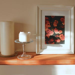 May include: A wooden shelf displays a framed floral print, a large pillar candle, and a small candle on a glass stand. The framed print features red and pink flowers against a dark background. The shelf is made of wood.