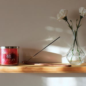 May include: A wooden shelf holds a red candle, incense, and a clear glass vase with white flowers. The candle has a silver lid and a label that reads "'Tis the Season". The incense is black, and the vase is filled with water.