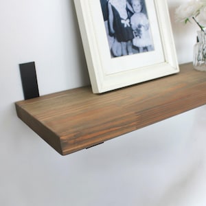 May include: A stained wooden floating shelf, supported by black metal brackets. A framed black and white photograph and a small glass vase with white flowers are displayed on the shelf. The shelf is mounted on a white wall.