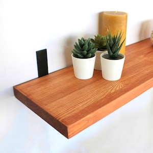 May include: A wooden floating shelf with a warm, reddish-brown finish, supported by black metal brackets. The shelf is decorated with small potted succulents, a tall yellow candle, and a woven basket with a lid. The shelf is approximately 76 cm long.